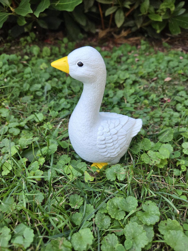 K&ouml;nnte beinhalten: Eine wei&szlig;e G&auml;nsefigur mit gelbem Schnabel und F&uuml;&szlig;en, vor gr&uuml;nem Gras und Laub. Die Gans hat eine detaillierte, strukturierte Oberfl&auml;che und ein schwarzes Auge, eine charmante Gartendekoration.