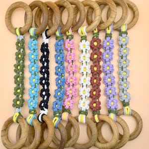 Puede incluir: Una colección de aros de madera con detalles florales de macramé de colores. Las flores son de varios colores, incluyendo azul, rosa, blanco y morado. Los aros están conectados por el macramé, creando una exhibición decorativa.