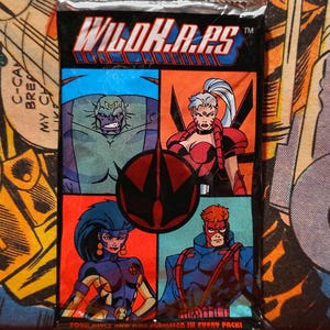 May include: A sealed trading card pack with the title "WILDK.A.PS" in bold letters. The pack features four comic-style illustrations of costumed characters in red, blue, and orange squares. The bottom of the pack reads "FOUR PIECE AND ONE HAMMER IN EVERY PACK!"