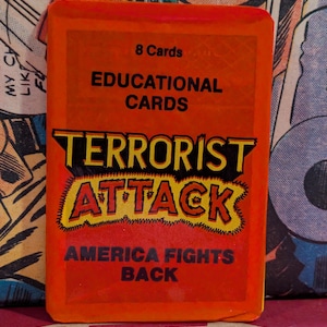 May include: An orange pack of "Terrorist Attack" educational cards. The text on the front reads "8 Cards", "Educational Cards", "Terrorist Attack", and "America Fights Back". The background is a comic book page.