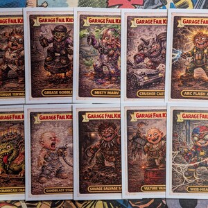 May include: A collection of vintage "Garage Fail Kids" trading cards. Each card features a grotesque cartoon character with a humorous name. The cards have a white border and a distressed, aged appearance, with titles like "Torque Tentacl" and "Grease Gobble".