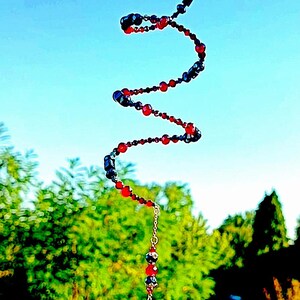 Könnte beinhalten: Ein hängendes Spiralornament mit einer Reihe abwechselnder roter und schwarzer Perlen, die auf einen Draht gefädelt sind. Die Spirale ist mit einer Metallkette verbunden, die zu einer kleinen, klaren Glasflasche in Totenkopfform mit Holzverschluss führt.