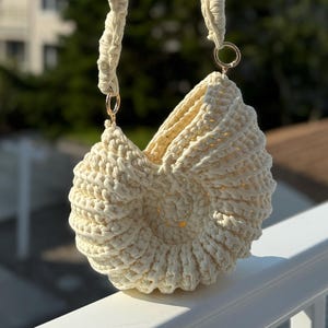 May include: Cream-colored, handmade crochet bag with a unique, shell-like design. The bag features a rounded shape, a textured surface, and a braided handle attached with gold-colored rings. The bag is held up against a backdrop of a sunny sky and houses.