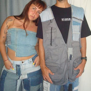 May include: A couple wearing patchwork denim clothing. The woman wears a denim tube top and patchwork jeans. The man wears a black t-shirt, a gray vest with denim patches, and patchwork jeans. Both are posing in front of a gray curtain.