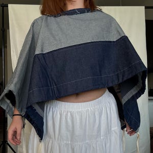 May include: A denim poncho in shades of gray and dark blue, paired with a white tiered skirt. The poncho has a raw edge design. The skirt has an elastic waistband. Cream-colored ties with metal beads hang from the poncho.