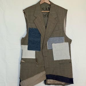 May include: A sleeveless, patchwork vest in shades of brown, beige, and blue. The vest is made from a suit jacket, with various fabric patches sewn onto it. The vest is hanging on a wooden hanger.