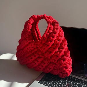 Peut inclure: Un sac à main rouge vif, tricoté en grosses mailles, avec une anse arrondie. Le sac est fait d'un fil épais et texturé. Le sac est posé sur un ordinateur portable.