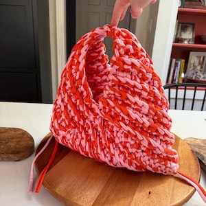 May include: A handmade, crocheted tote bag in shades of red and pink. The bag features a unique, open handle design and is made with thick, textured yarn. The bag is sitting on a wooden cutting board.