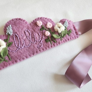 May include: A lilac felt crown embroidered with the name "Lily". The crown is decorated with embroidered flowers in white, pink, and blue, with green leaves. A matching lilac satin ribbon is attached for tying.