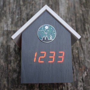 Puede incluir: Un reloj de madera gris oscuro con forma de casa con una luna plateada llena de estrellas y dos figuras mirando a través de un telescopio. La esfera del reloj muestra el número 1233 en rojo.