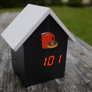 K&ouml;nnte beinhalten: Eine schwarze, hausf&ouml;rmige Uhr aus Holz mit einem roten digitalen Display, das die Zeit 10:11 anzeigt. Auf der Vorderseite der Uhr befinden sich eine rote Kaffeetasse und ein Donut mit Streuseln.