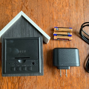 K&ouml;nnte beinhalten: Eine schwarz-wei&szlig;e hausf&ouml;rmige Uhr mit digitaler Anzeige und Kn&ouml;pfen zum Einstellen der Zeit. Die Uhr wird mit drei AA-Batterien oder einem Netzteil mit USB-Kabel betrieben. Die Uhr hat eine kleine &Ouml;ffnung an der Vorderseite zum Einstellen der Zeit.