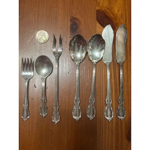 May include: A collection of antique silverware, including forks, spoons, and knives, arranged on a wooden surface. The silverware has ornate handles and a reflective, silver finish. A US quarter is also present.