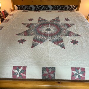 May include: A large, cream-colored quilt with a central starburst design in shades of red, gray, and white. Smaller star patterns are scattered across the surface. The quilt is placed on a wooden bed frame.