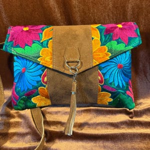 May include: A colorful embroidered clutch with a brown suede flap and tassel. The clutch features vibrant floral patterns in pink, blue, yellow, and green. The bag has a brown strap.