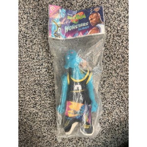 May include: A blue Monstars basketball player toy in a clear plastic bag. The toy is wearing a black and yellow jersey with the number 5. The packaging features the Monstars logo and the words "Monstars Blanko".