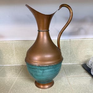 May include: A tall pitcher featuring a metallic copper upper section and a teal-colored lower base. The vessel has a curved handle, a flared spout, and a decorative twisted rope-like band separating the copper and teal portions of the body.