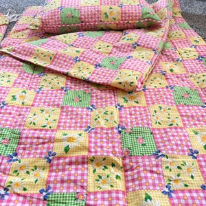 May include: A quilted bedding set featuring a patchwork pattern of pink gingham, green gingham, and yellow polka dot squares. The fabric is decorated with white daisy and blue flower prints throughout the quilt and the matching rectangular pillow.