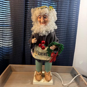 May include: A whimsical Christmas figurine of an elderly man with a white beard and curly hair, wearing a dark coat and green pants. He holds a small wreath and a bag that reads "NAILS SCREWS". The figurine has brown shoes and a smiling face.