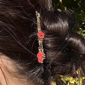 May include: A gold-colored hair pin with a chain and three red flower charms. The hair accessory is inserted into dark brown hair, with the flowers dangling down. The hair is styled in a bun.