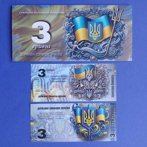 Op de afbeelding: Drie Oekraïense hryvnia souvenirbiljetten. De bankbiljetten tonen het Oekraïense wapen, een drietand en de Oekraïense vlag in blauw en geel. Het cijfer "3" staat prominent op elke biljet.