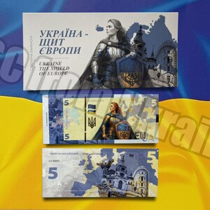 Puede incluir: Una colección de billetes ucranianos y una tarjeta conmemorativa. La tarjeta presenta a una mujer con armadura y un escudo, con el texto "Ukraine The Shield of Europe". Los billetes son azules, amarillos y blancos, con el número "5".