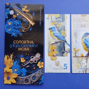 Op de afbeelding: Een verzameling Oekraïense souvenirbiljetten en een boekje. De bankbiljetten tonen een blauw-gele vogelillustratie, met het nummer 5. Het boekje heeft de tekst "SOLOV'INA UKRAINSKA MOBA" en bloemmotieven.