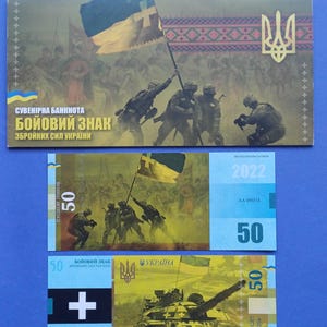 Op de afbeelding: Drie Oekraïense souvenirbiljetten op een blauwe achtergrond. De bovenste biljet toont soldaten die een vlag hijsen, met Oekraïense tekst. De andere twee biljetten tonen een tank en een vlag, met het getal 50.