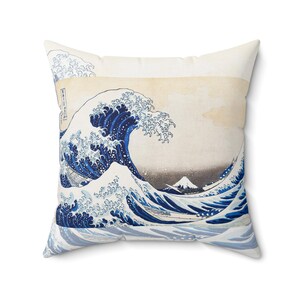 以下が含まれることがあります： 日本の象徴的な芸術作品「神奈川沖浪裏」のプリントが施された正方形のクッション。デザインは、青色の濃淡で表現された大きく様式化された波と、背景に富士山が描かれています。クッションは白い背景です。