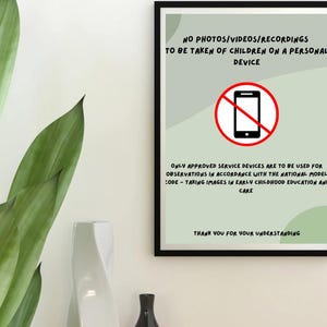 May include: A framed poster with a black border displays text in black font on a light green background. The text reads "NO PHOTOS/VIDEOS/RECORDINGS TO BE TAKEN OF CHILDREN ON A PERSONAL DEVICE." A red circle with a line through a smartphone graphic is in the center.
