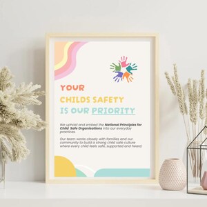 May include: A framed print with the text "YOUR CHILDS SAFETY IS OUR PRIORITY." The print features colorful handprints and pastel design elements. The print is displayed on a white surface with decorative vases and dried plants.