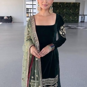 Könnte beinhalten: Ein dunkelgrüner Salwar Kameez aus Samt mit goldener Stickerei an Ärmeln und Saum. Das Outfit beinhaltet ein durchsichtiges, hellgrünes Dupatta mit goldenen Verzierungen und einem bordeauxfarbenen Rand. Der quadratische Ausschnitt und die verzierten Details schaffen einen eleganten Look.