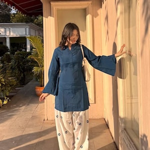 May include: A dark teal tunic top with flared sleeves and white pants with a black eye pattern. The outfit is a casual style, suitable for everyday wear. The ensemble is paired with matching shoes and a white shoulder bag.