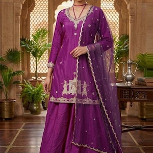 Puede incluir: Un vibrante conjunto de salwar kameez púrpura. El atuendo presenta una túnica de manga larga con intrincados bordados dorados, combinada con pantalones de pierna ancha a juego y una dupatta transparente. El atuendo está adornado con ribetes y adornos dorados.