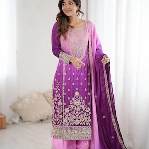 May include: A vibrant, embroidered salwar kameez in shades of purple and pink. The long-sleeved tunic features intricate gold detailing, paired with matching palazzo pants and a flowing dupatta. The ensemble is a traditional South Asian outfit.
