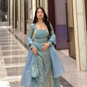 Puede incluir: Un vestido formal azul claro con un chal y un bolso a juego. El vestido presenta un escote cuadrado y mangas largas. La tela tiene un brillo sutil y el atuendo está adornado con delicados bordados. El bolso es una pequeña bolsa con cordón.
