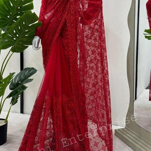 Könnte beinhalten: Ein leuchtend roter Sari mit aufwendigen Pailletten- und Spitzendetails. Der Sari hat einen transparenten, gemusterten Überzug und eine einfarbige rote Unterschicht. Die langen Ärmel und das drapierte Design erzeugen eine elegante Silhouette. Eine weiße Halskette ist sichtbar.
