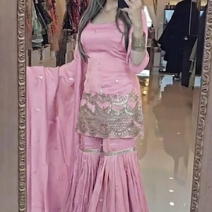 Könnte beinhalten: Ein hellrosa, dreiteiliges Ensemble mit einem quadratischen Ausschnitt, ausgestellten Hosen und einem passenden Schal. Das Outfit ist mit aufwendiger goldener Stickerei am Saum und an den Manschetten verziert. Es ist ein traditioneller südasianischer Stil.