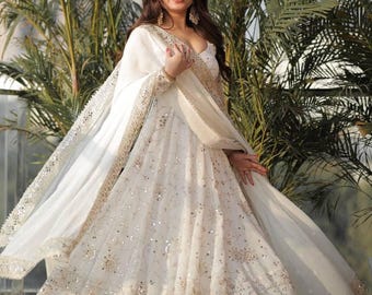 May include: A white, flowing dress with intricate sequin and embroidery details. The dress features a deep V-neckline and is paired with a matching shawl. The edges of the shawl are embellished with a delicate trim.