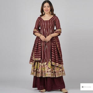 Puede incluir: Un vestido Anarkali color granate con escote redondo, mangas tres cuartos y un corpiño con rayas verticales blancas y detalles dorados. La falda presenta un estampado floral grande en tonos verde y rosa apagados, combinado con pantalones anchos a juego.