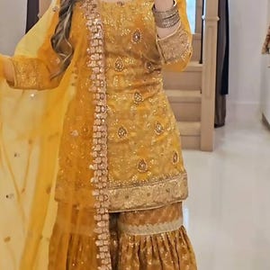 Puede incluir: Traje Sharara bordado en amarillo mostaza. El atuendo presenta intrincados bordados y adornos dorados. El conjunto incluye una túnica de manga larga, pantalones de pernera ancha y un dupatta transparente. El atuendo está adornado con lentejuelas y ribetes dorados.