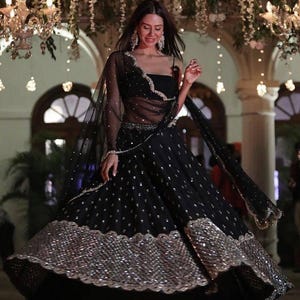 Puede incluir: Un vestido de noche negro con una parte superior transparente de manga larga y una falda amplia. La falda está adornada con lentejuelas plateadas y un borde ancho y brillante. El vestido se complementa con pendientes colgantes y una pulsera delicada.