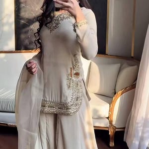 May include: A beige embroidered formal outfit. The ensemble features a long-sleeved tunic with gold embroidery, wide-leg trousers, and a matching sheer scarf. The outfit is suitable for special occasions and events.