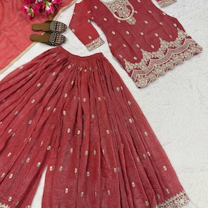May include: A red embroidered Indian suit set, including a long-sleeved top, wide-leg pants, and a matching scarf. The top and pants feature intricate gold embroidery along the neckline, cuffs, and hem. A pair of brown sandals with decorative beading are also present.