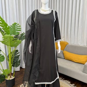 Puede incluir: Un conjunto negro de tres piezas que consta de una túnica de manga larga, pantalones anchos y una dupatta de organza transparente. La túnica presenta un cuello redondo y ribetes de encaje blanco en los bordes. Se lleva un collar de perlas blancas.
