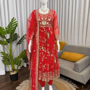 Puede incluir: Un vibrante conjunto formal rojo, con una túnica de manga larga y pantalones a juego, adornado con intrincados bordados dorados y detalles de lentejuelas. Una bufanda roja transparente cae elegantemente. El atuendo se exhibe en un maniquí.
