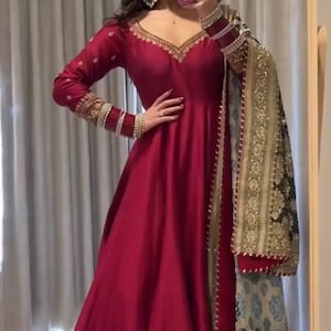 Puede incluir: Un vestido largo y fluido de color rojo con mangas largas y bordados dorados en los puños y el escote. Una chal a juego con detalles dorados y negros está drapeado sobre el hombro. El vestido tiene un escote en V.