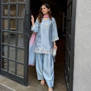 Puede incluir: Conjunto salwar kameez de seda azul claro. La parte superior presenta detalles bordados alrededor del escote y las mangas, con bordados a juego en el dobladillo del pantalón. Una bufanda degradada rosa y blanca y un clutch plateado completan el atuendo.
