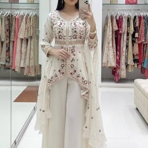 Peut inclure: Ensemble tunique et pantalon palazzo brodé, de couleur ivoire. La tunique présente un col en V et des broderies florales rouges et marron. Le pantalon palazzo est à jambes larges. L'ensemble est présenté dans une boutique de vêtements.