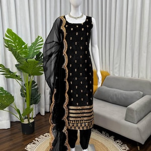 Puede incluir: Traje sin mangas de terciopelo negro con bordados dorados y una bufanda transparente a juego. El traje presenta intrincados detalles dorados en el corpiño, el dobladillo y los bordes de la bufanda. El conjunto se exhibe en un maniquí.